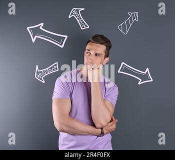 Choice in profession or other areas of life, concept. Making decision, thoughtful young man surrounded by drawn arrows on grey background Stock Photo