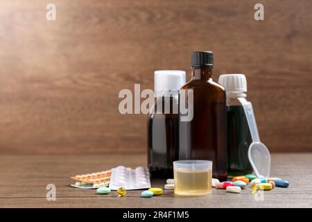 Bottles of syrup, measuring cup, dosing spoon and pills on wooden table, space for text. Cold medicine Stock Photo