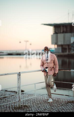 Vertical shot, an outdoor scene with pink skies. Tagus River backdrop, mature bald black man with long white-bearded, exuding elegance in pink wrap ja Stock Photo