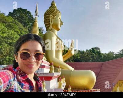 A young Thai woman taking a selfie in front of a Buddha statue at Wat Khao Rang, a Buddhist temple in Phuket, Thailand. Stock Photo