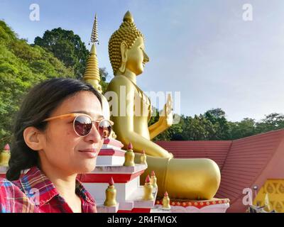 A young Thai woman taking a selfie in front of a Buddha statue at Wat Khao Rang, a Buddhist temple in Phuket, Thailand. Stock Photo
