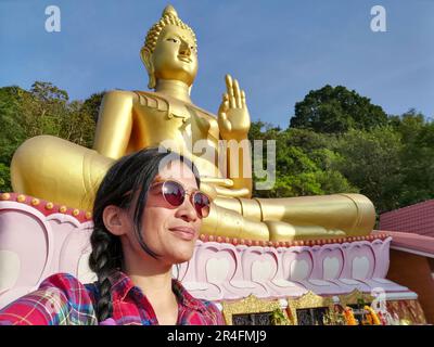 A young Thai woman taking a selfie in front of a Buddha statue at Wat Khao Rang, a Buddhist temple in Phuket, Thailand. Stock Photo