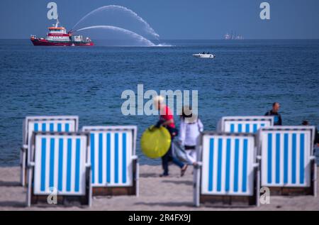 Rostock, Germany. 28th May, 2023. The fireboat 'Albert Wegener' sprays water while the first vacationers walk past the mostly still empty beach chairs on the Baltic coast in the morning. With lots of sunshine and high temperatures, the weather shows its best side on Whit Sunday in northern Germany. Credit: Jens Büttner/dpa/Alamy Live News Stock Photo