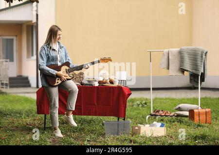 Woman holding guitar near table with different items on garage sale in yard Stock Photo