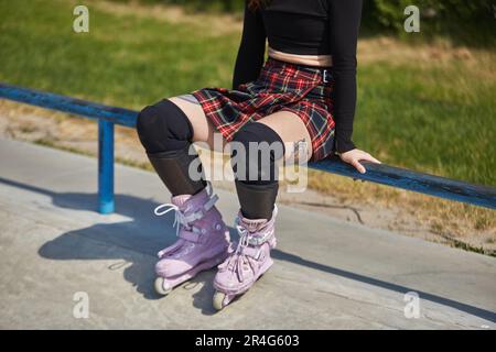 Tattooed roller blader girl sitting on a rail in a skatepark. Cool young female athlete wearing modern aggressive inline skates and protective knee pa Stock Photo
