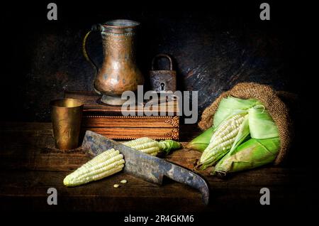 Still life with raw organic white corns placed in burlap bag with vintage jar, old copper box and old cleaver on rustic dark background.. Stock Photo