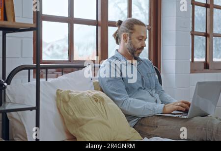 Young worried man using laptop sitting on bed at bedroom. Stock Photo