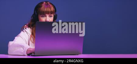 Young female gamer looking at her laptop screen while playing an online video game in a studio. Woman with ginger hair using an interactive streaming Stock Photo