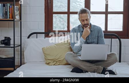 Young worried man using laptop sitting on bed at bedroom. Stock Photo