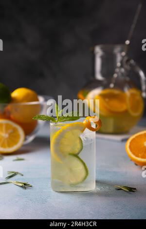 Refreshing cold summer drink with orange, lemon, lime, mint and rosemary. A misted glass with ice and citrus slices, in the background a jug with a le Stock Photo