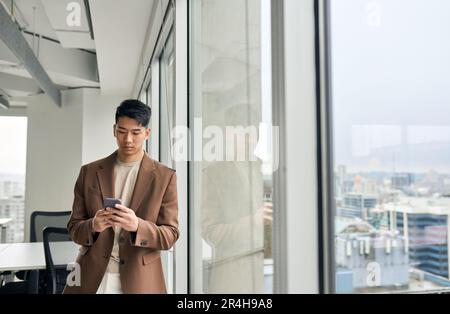Young Asian business man using smartphone standing in office at window. Stock Photo