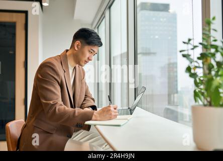 Young busy Asian business man working on laptop in office writing notes. Stock Photo