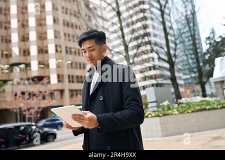 Young smiling Asian business man walking in city using digital tablet. Stock Photo