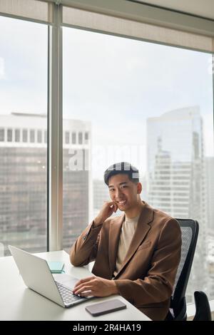 Young happy Asian businessman looking at camera working on laptop in office. Stock Photo