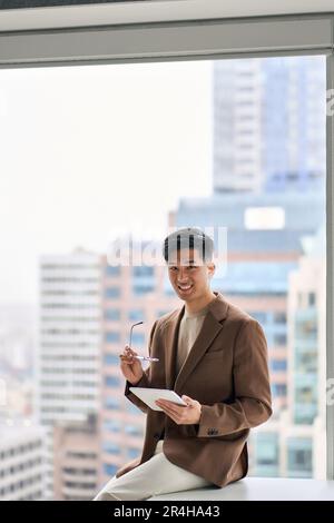 Young happy Asian business man holding tablet sitting in office. Vertical Stock Photo