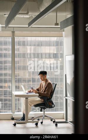 Young Asian business man using phone in office sitting at desk, vertical. Stock Photo