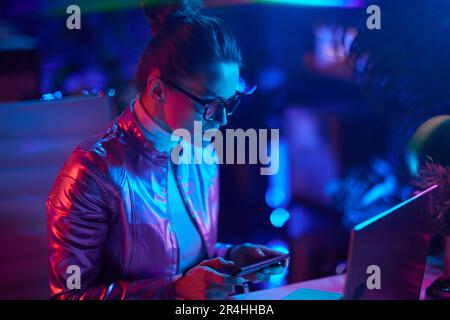 Neon metaverse futuristic concept. elegant female in glasses with laptop using smartphone in modern office. Stock Photo