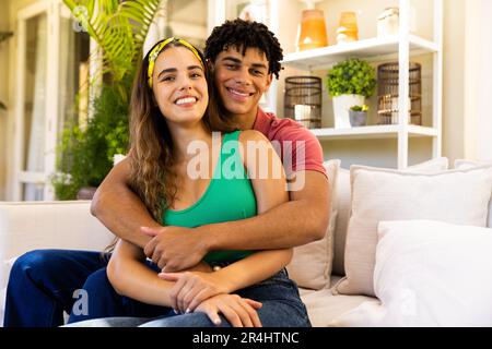 Portrait of smiling biracial young man embracing girlfriend while sitting on sofa in living room Stock Photo