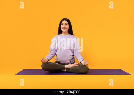 Peaceful expectations. Happy young pregnant lady sitting on yoga mat, meditating and exercising over yellow background Stock Photo