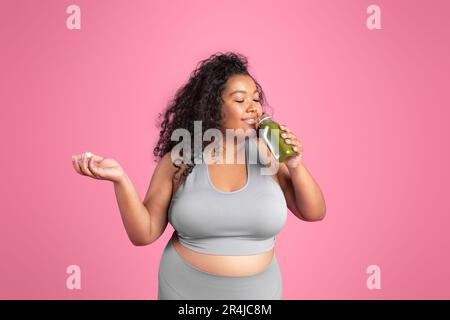 Positive black plus size woman in sportswear holding jar of fresh smoothie, standing on pink studio background Stock Photo
