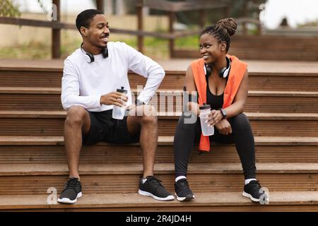Cheerful young black couple sitting on stairs, talking, relaxing with bottles of water in summer Stock Photo