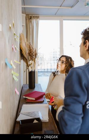 Male and female colleagues discussing over sticky notes on wall in creative office Stock Photo