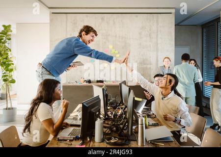Cheerful entrepreneurs giving high-five while colleagues sitting at desk in creative office Stock Photo