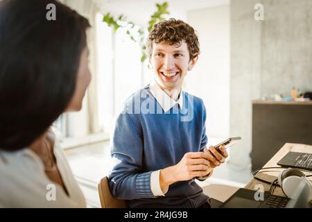 Happy young entrepreneur holding smart phone discussing with colleague in creative office Stock Photo