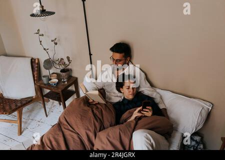 High angle view of man reading book by non-binary person using mobile phone on bed at home Stock Photo