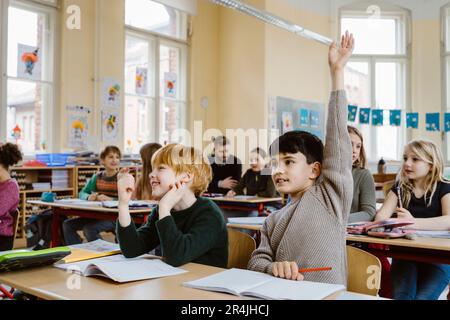 Boy with hand raised answering during lecture while sitting by male friend in classroom Stock Photo