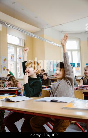 Smiling male pupils raising hands while answering in classroom Stock Photo