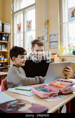 Happy male teacher assisting schoolboy using tablet PC at desk in school Stock Photo
