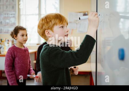 Side view of blond boy writing on whiteboard while solving mathematics in classroom Stock Photo