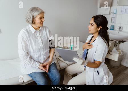 Female doctor holding tablet PC advising senior patient sitting on bed in clinic Stock Photo