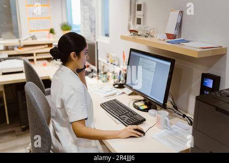 Female healthcare worker using computer while talking on smart phone at clinic Stock Photo