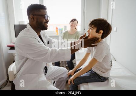 Smiling male pediatrician examining neck of boy sitting on bed at healthcare center Stock Photo