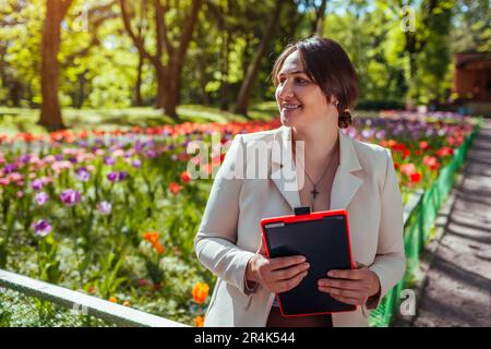 Portrait of happy businesswoman holding tablet sitting in spring park. Female landscape designer enjoys results of blooming flowerbeds in public garde Stock Photo