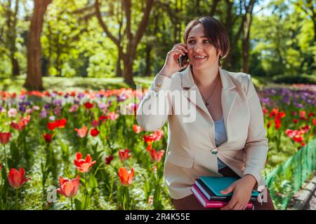 Young caucasian businesswoman talking on phone holding notebooks in spring park. Teacher having break outdoors relaxing in university campus. Stock Photo