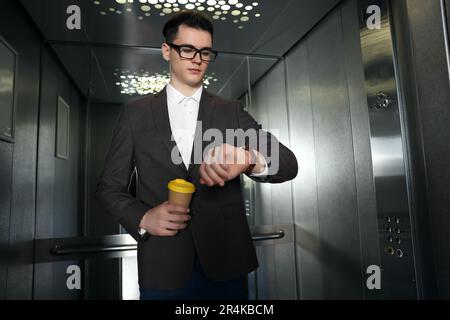 Young businessman with cup of hot drink checking time in elevator Stock Photo