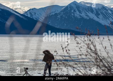 Man standing beside partial frozen thawing lake in Canada during spring time with snow capped mountains in background in wilderness area. Stock Photo