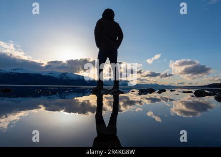 Unique view of a person with reflection in a natural lake with clouds and mountains in background. Awesome perspective of person. Stock Photo