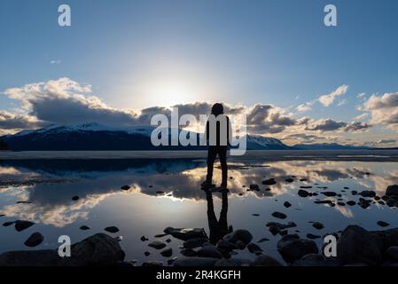 Unique view of a person with reflection in a natural lake with clouds and mountains in background. Awesome perspective of person. Stock Photo