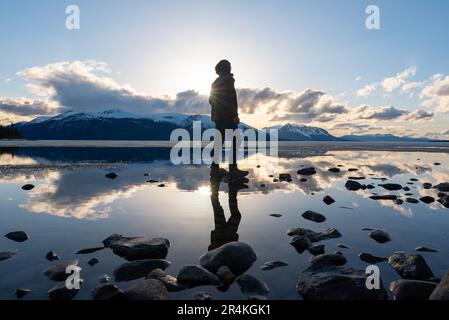 Unique view of a person with reflection in a natural lake with clouds and mountains in background. Awesome perspective of person. Stock Photo