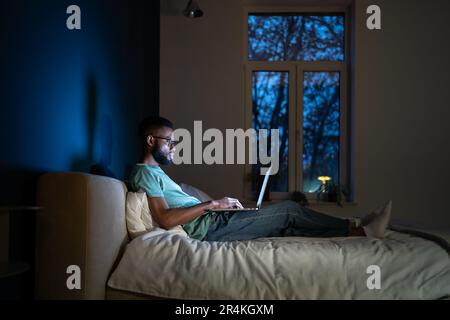 Concentrated African American programmer man lying on bed work on laptop at night.  Stock Photo