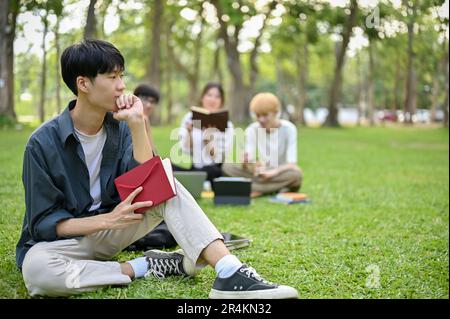 A thoughtful young Asian male college student sits on the grass in the campus park with his book, thinking, pondering, or planning something while loo Stock Photo