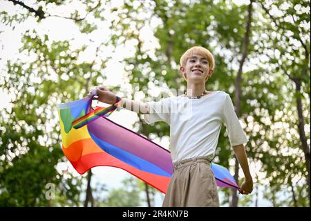 Portrait of an attractive and carefree young Asian gay man with an LGBT rainbow flag standing in the green park. Diversity and human rights, Pride mon Stock Photo