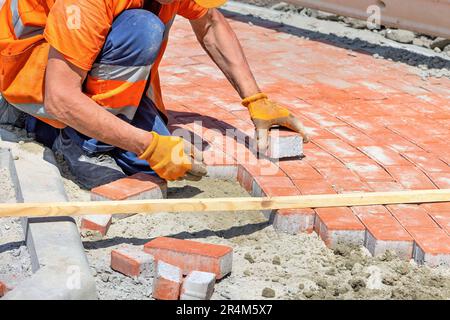 A bricklayer in work gloves and overalls lays red paving slabs on a sunny day. Stock Photo