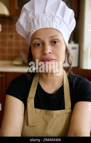 Confident portrait of a beautiful female baker confectioner, wearing white chef's hat and beige apron, looking at camera Stock Photo