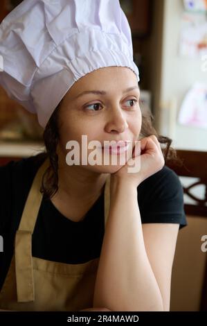 Authentic portrait of a beautiful female confectioner, wearing white chef's hat and beige apron, dreamily looking aside Stock Photo