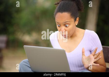Surprised black woman checking laptop content sitting on a bench in a park Stock Photo